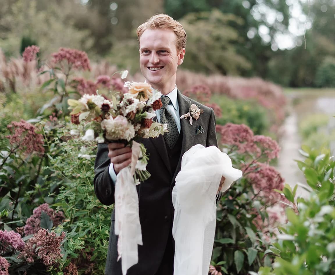 Groom wearing a bespoke suit holds bride's bouquet and veil