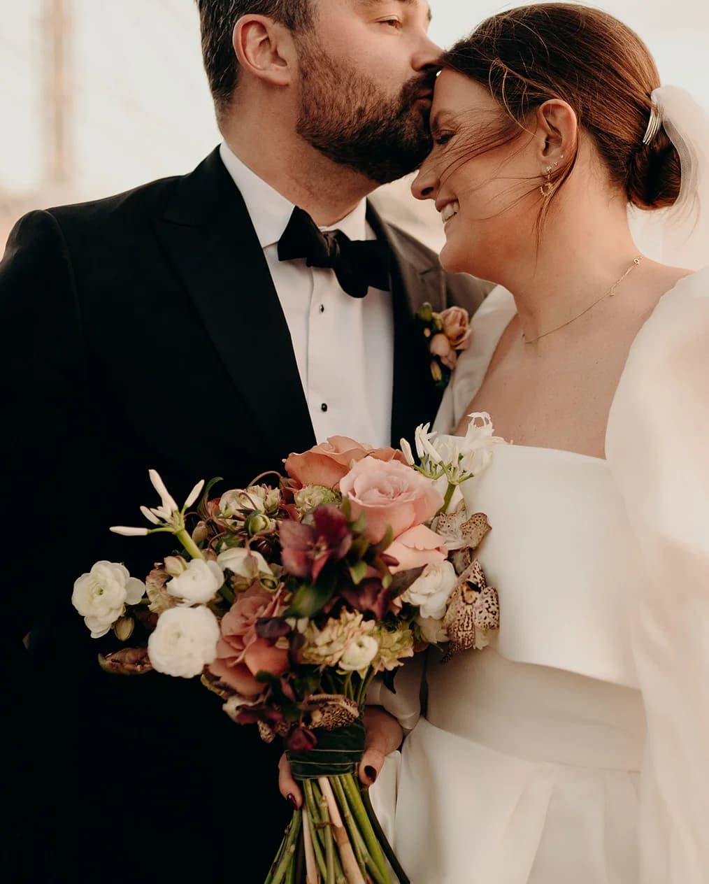 Groom kisses bride on the forehead, while she is holding winter flower bouquet