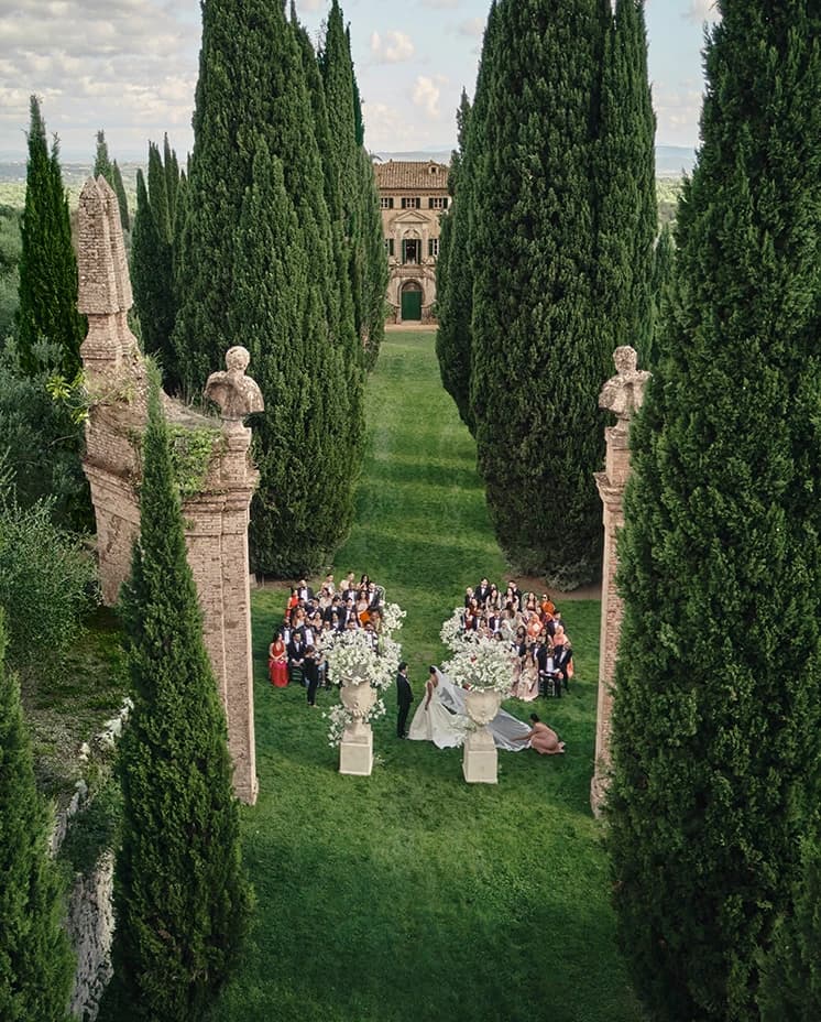 Elegant outdoor destination wedding ceremony in Villa Cetinale’s garden, set along a cypress-lined avenue at a historic Italian villa, with guests gathered around the couple beneath grand stone urns.
