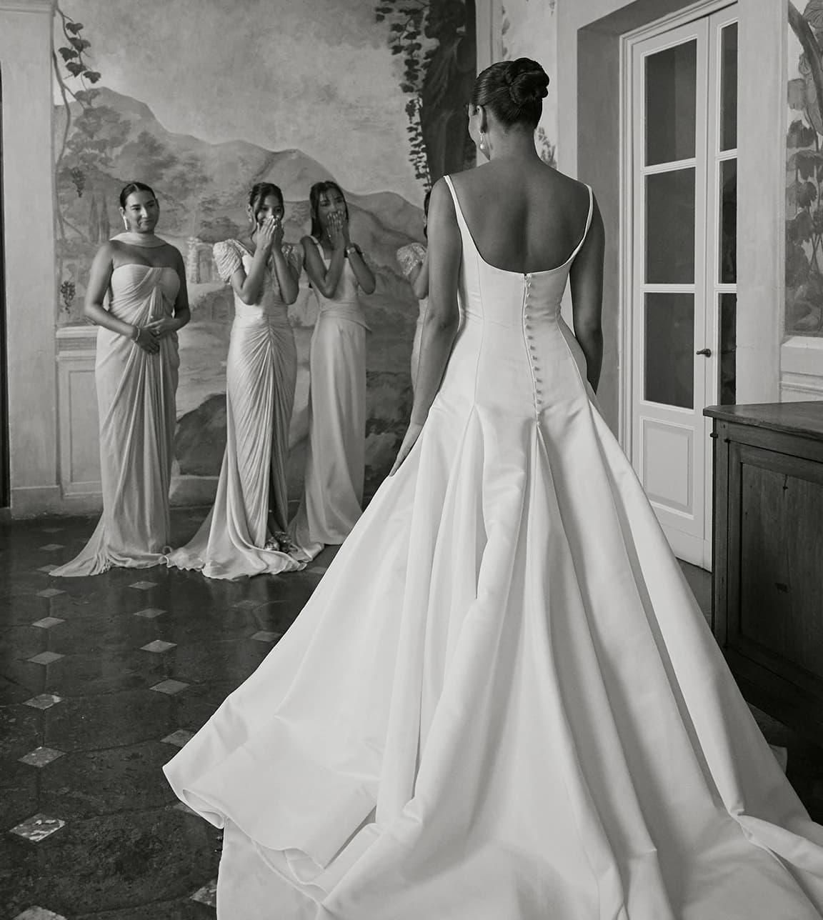 A bridal moment featuring bridesmaids in elegant, draped gowns reacting to the bride
