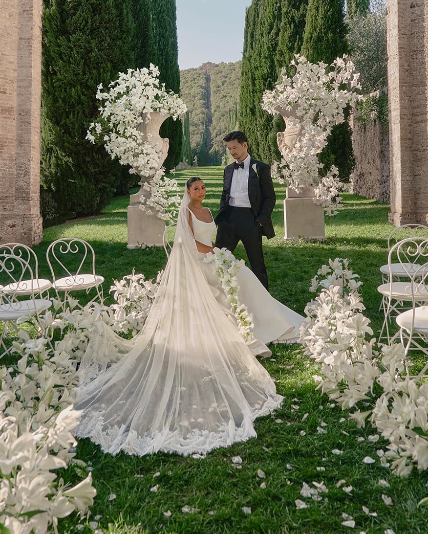 Elegant outdoor wedding setting at Villa Cetinale, with a cypress-lined aisle adorned with abundant white lilies and grand floral urn arrangements surrounding the couple