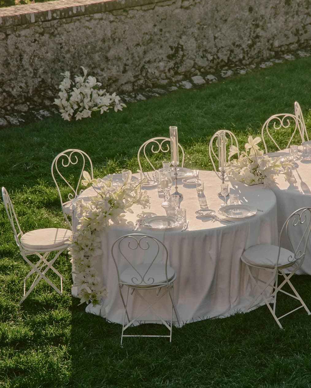 Elegant outdoor wedding tablescape with white linens, delicate glassware, and cascading white floral arrangements, set on a sunlit lawn