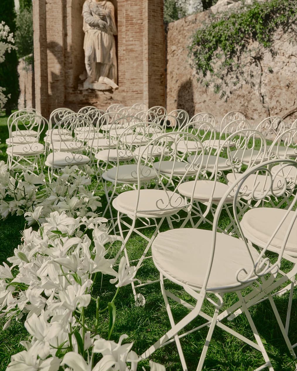Outdoor wedding ceremony setup with elegant white chairs and a lily-lined aisle, set against historic stone architecture and lush greenery