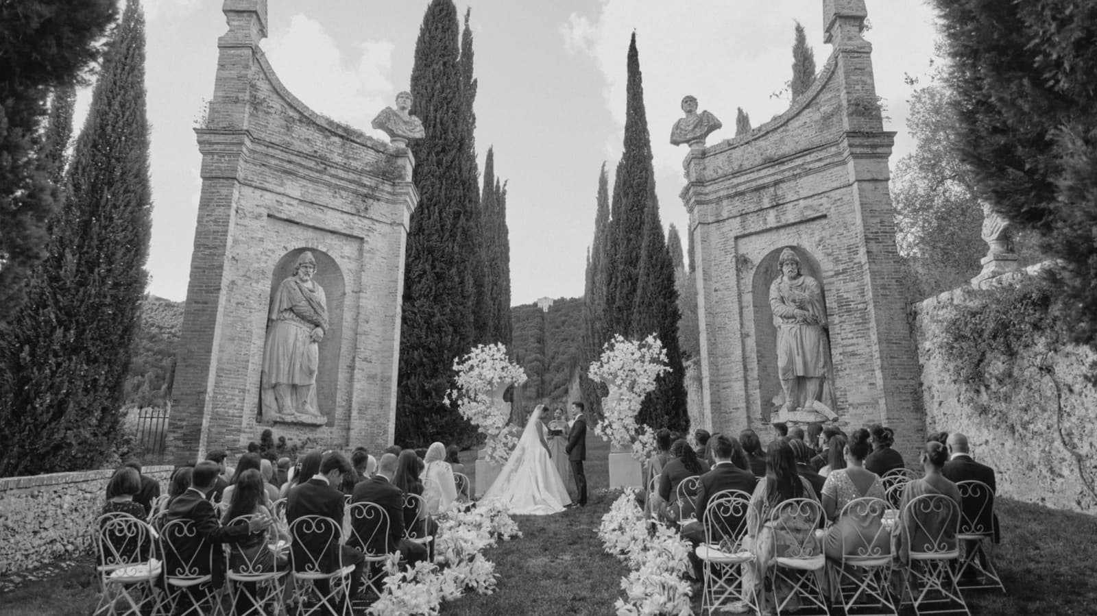 Black-and-white outdoor wedding ceremony at an Italian villa, with guests seated along a flower-lined aisle beneath cypress trees and historic stone arches.  Select 68 more words to run Humanizer.