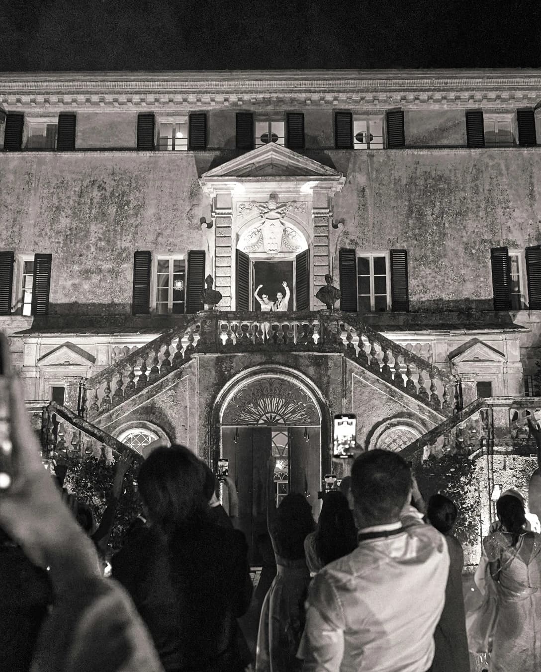 An evening scene of a historic Italian villa, with guests gathered below as the couple celebrates from a grand balcony.