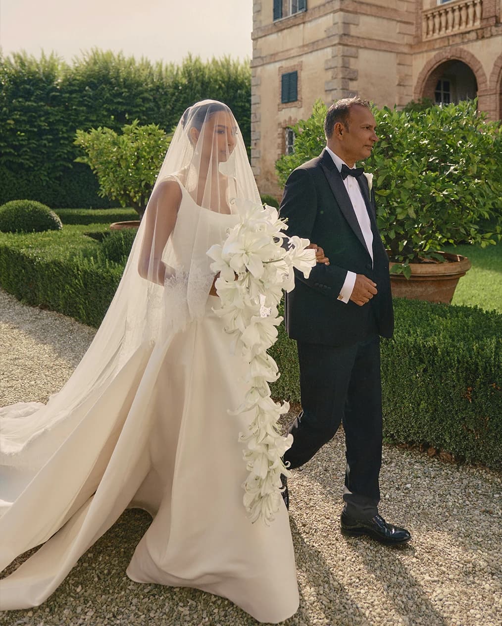 Bride walking through an Italian villa garden with a cascading bouquet of white lilies, accompanied by her father
