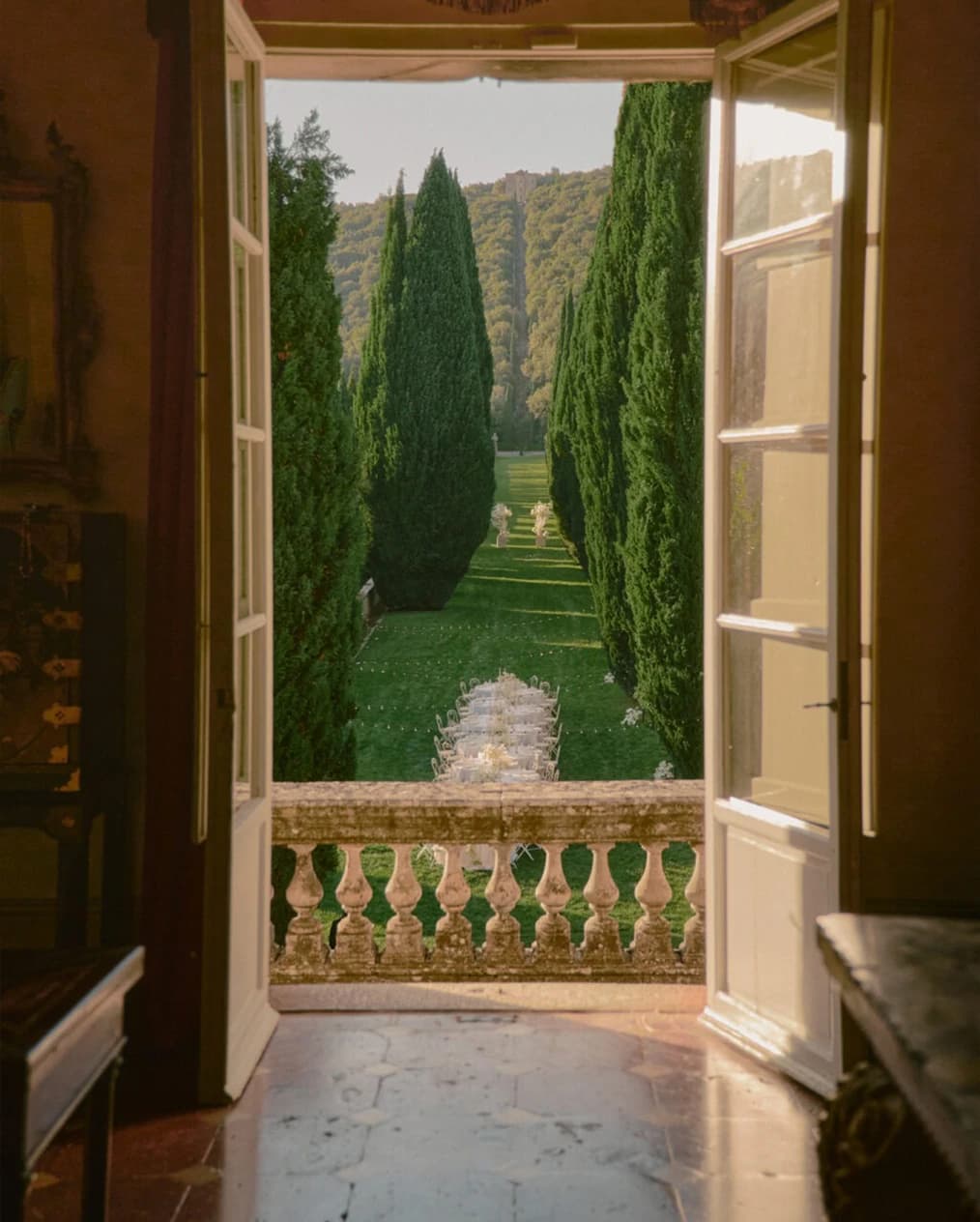View through open villa doors at Villa Cetinale, overlooking a cypress-lined garden aisle and elegant outdoor wedding setup bathed in warm light