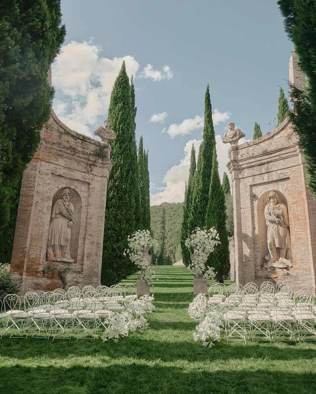 Outdoor wedding ceremony setup at Villa Cetinale, with a cypress-lined aisle, white chairs, and floral arrangements framed by historic stone arches