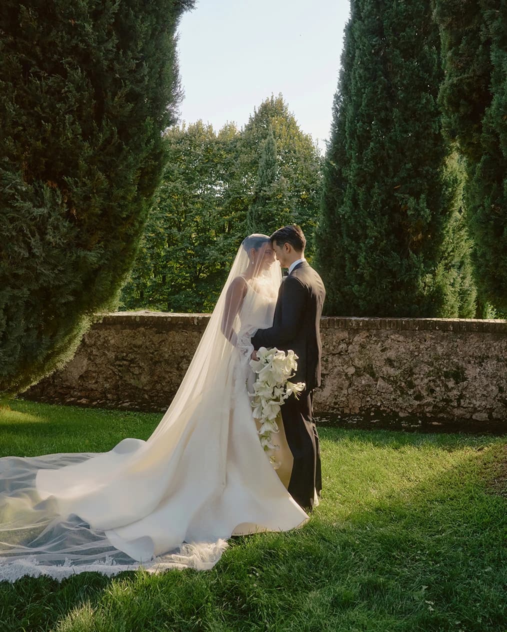 Romantic garden portrait at Villa Cetinale, with the couple standing beneath cypress trees, the bride holding a cascading bouquet of white lilies in soft golden light