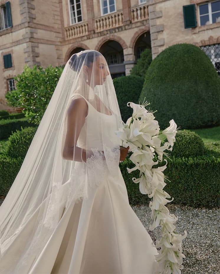 Bride walking through a villa garden during her wedding day, holding a cascading bouquet of white lilies beneath a sheer veil