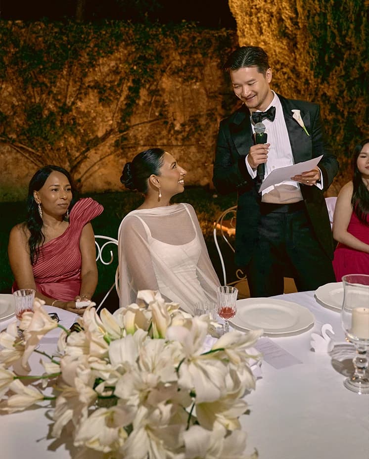 Wedding reception speech moment with the groom addressing guests, as the bride smiles beside him at a candlelit table adorned with white florals