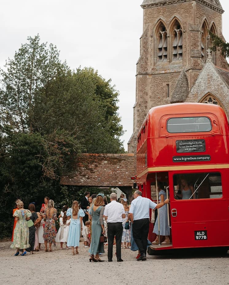 Wedding guests boarding a vintage red London double-decker bus outside a historic countryside church after the wedding ceremony.