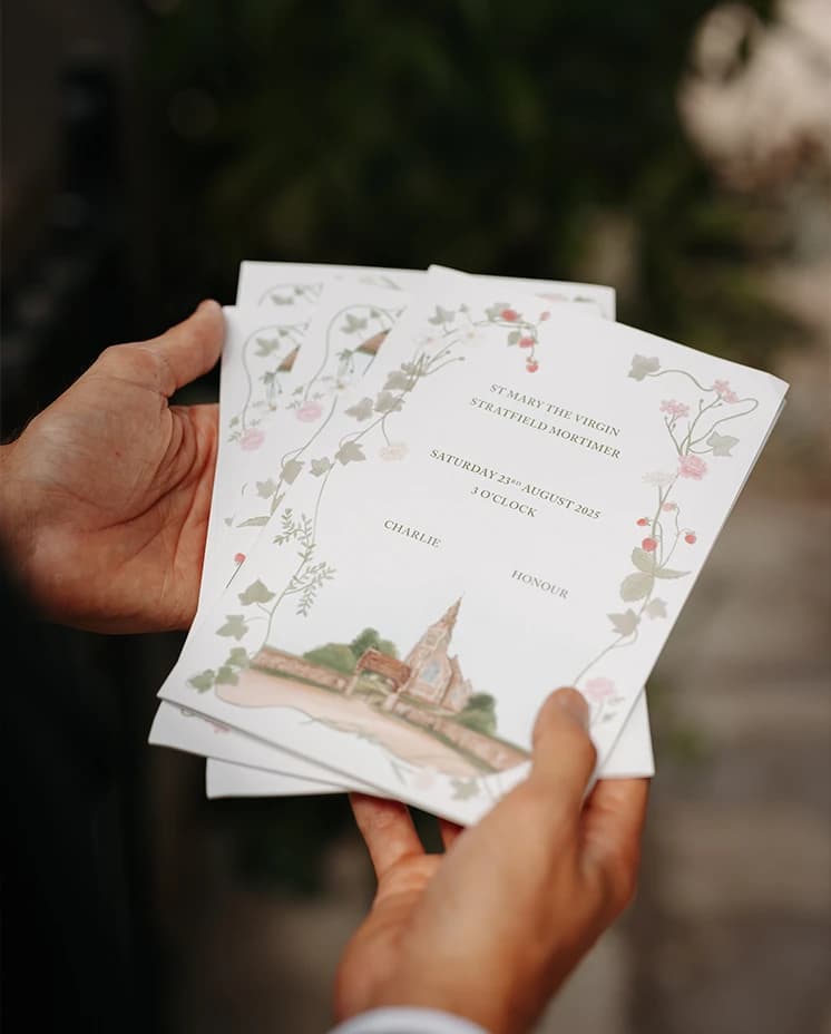 Hands holding an illustrated wedding order of service decorated with floral artwork and a painting of the ceremony church.