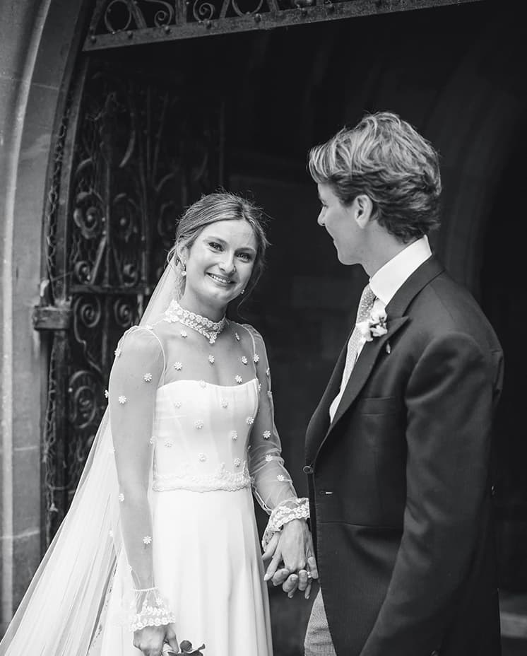 Black and white photograph of a Honour and Charlie holding hands and smiling outside a church after their wedding ceremony.