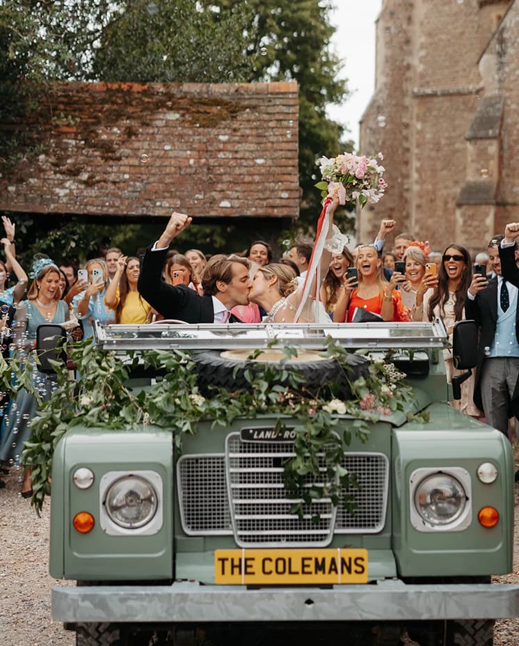 Bride and groom kissing in a vintage Land Rover adorned with greenery and a bespoke 'The Colemans' number plate, surrounded by cheering wedding guests outside a historic church.