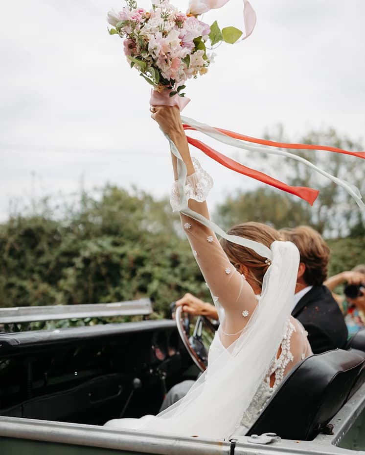 Bride holding her bouquet high with trailing ribbons as she rides in a vintage wedding car after the ceremony.
