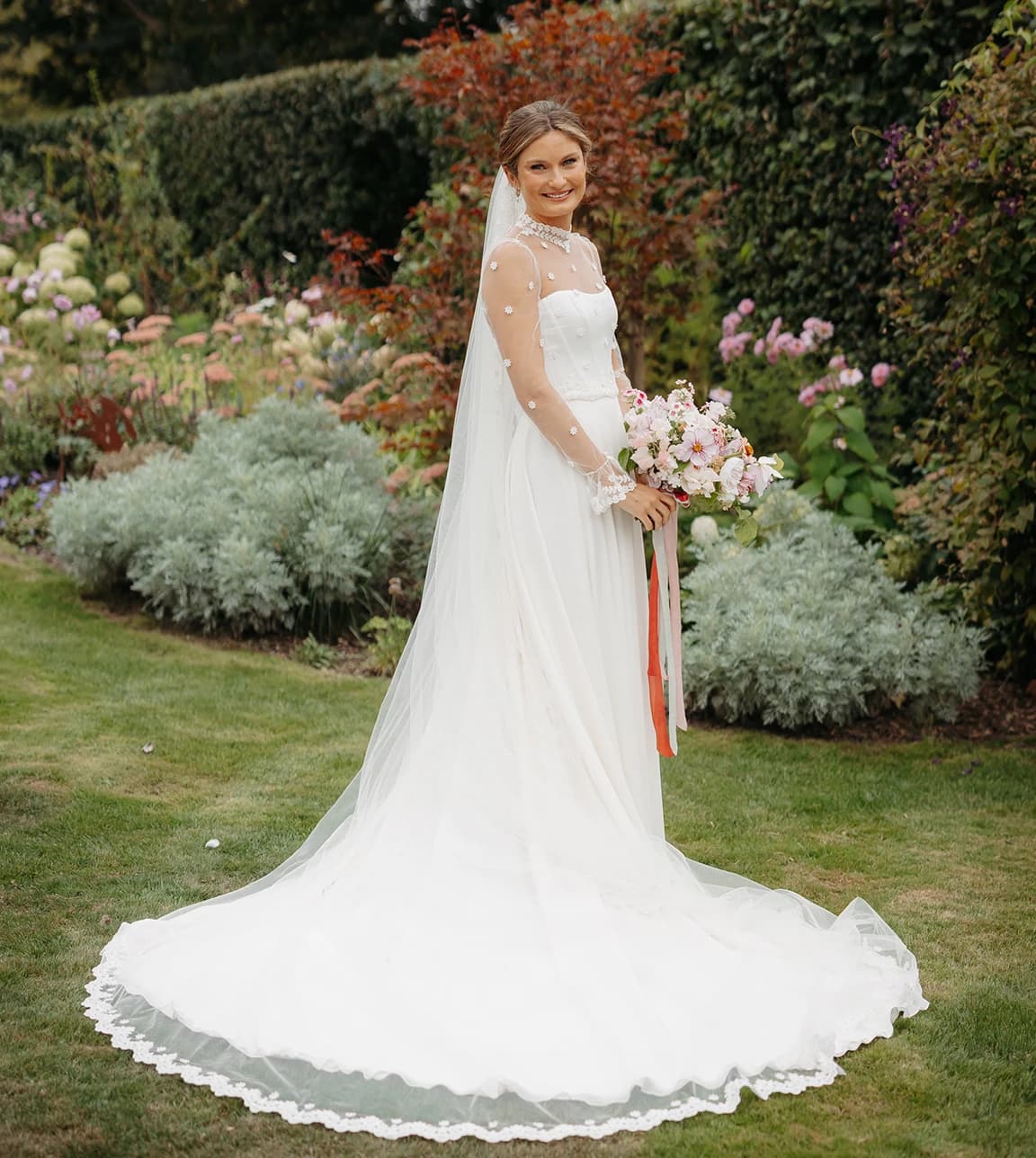 Honour standing in a garden holding a pastel wedding bouquet, wearing a flowing white gown and veil.