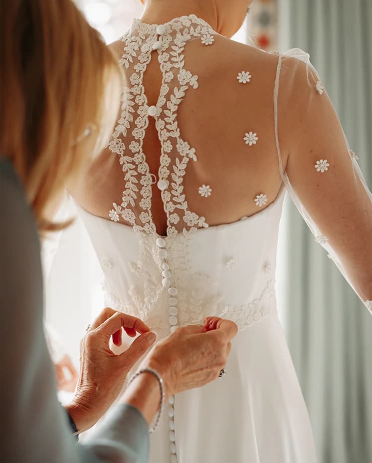 Bride having the buttons fastened on her lace-back wedding gown during morning preparations before a luxury countryside wedding.