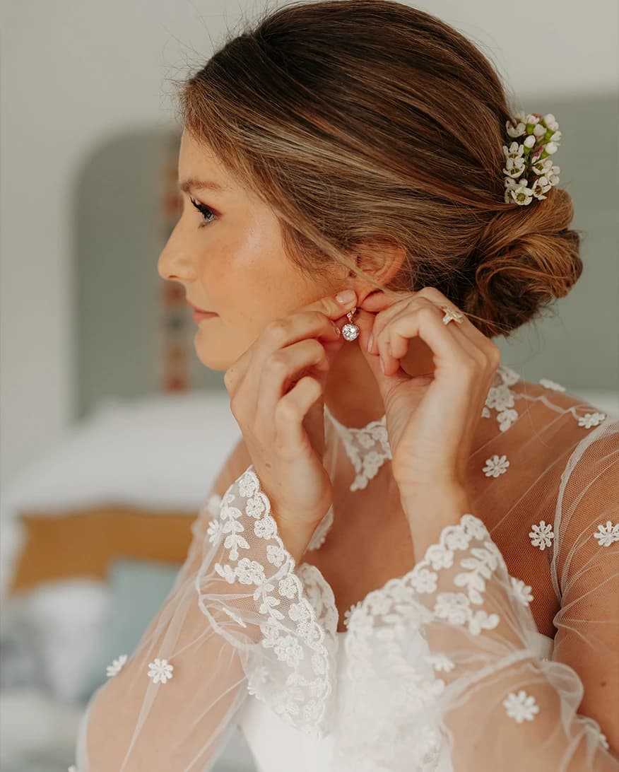 Bride putting on her Boodles earrings before the ceremony, featuring a romantic bridal updo adorned with delicate flowers and a lace wedding dress.