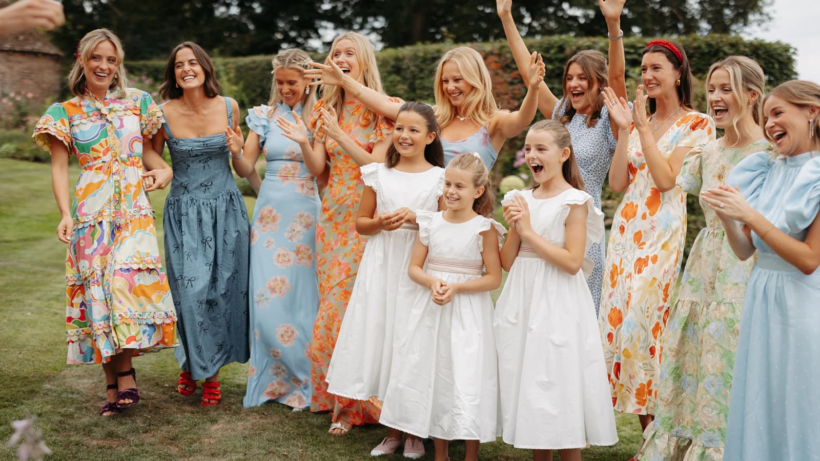 Bridesmaids wearing a mix of colourful floral-print and pastel blue dresses celebrate with flower girls in classic white dresses during a countryside wedding weekend.
