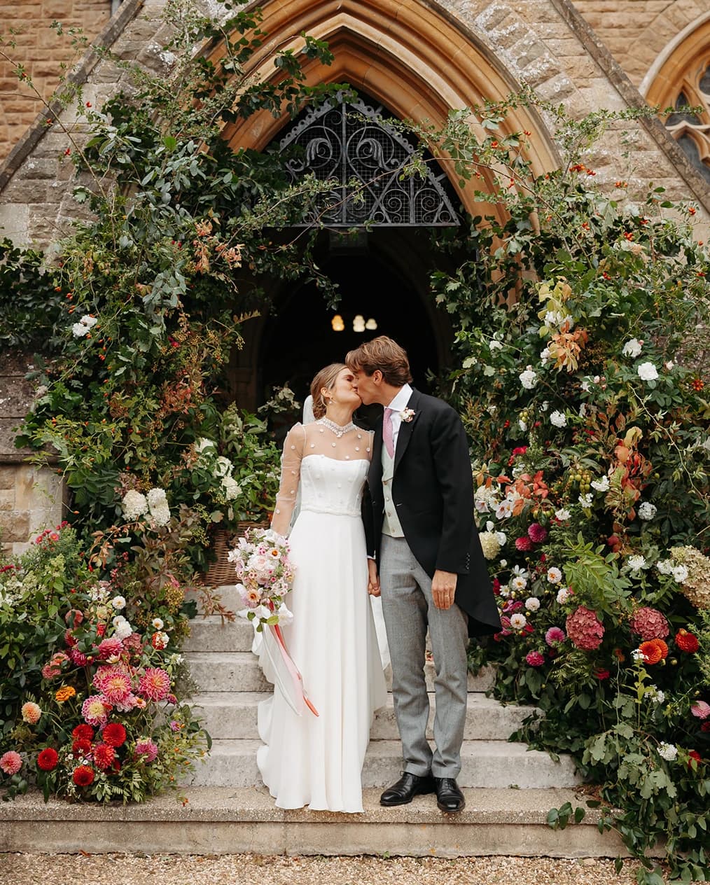 Bride and groom kissing beneath a breathtaking floral installation of seasonal blooms and cascading greenery at the entrance of a historic church.