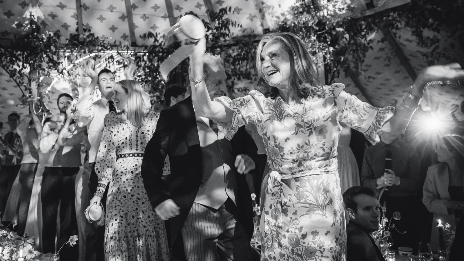 Wedding guests dancing with tambourines beneath cascading greenery and candlelit décor inside a luxury wedding marquee during the evening reception.