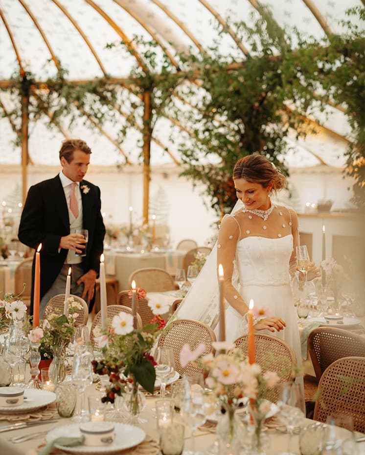 Bride and groom admiring their wedding reception décor inside a sailcloth marquee with candlelit tables, seasonal flowers and cascading greenery