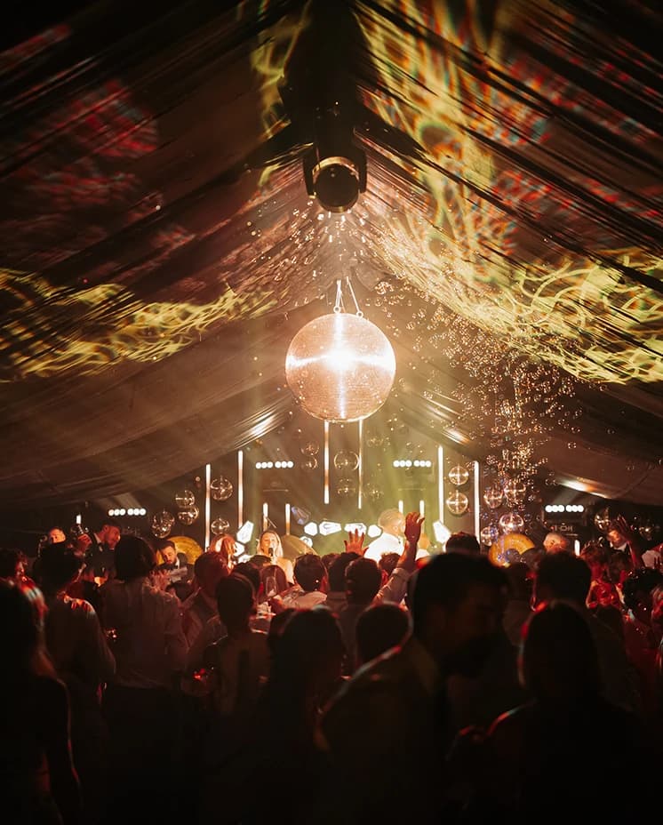 Packed dance floor inside a  wedding marquee illuminated by a sparkling disco ball and dramatic party lighting.