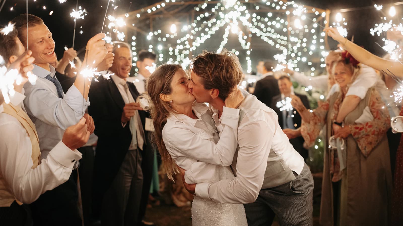 Newlyweds kissing during a magical sparkler send-off, surrounded by cheering guests and twinkling lights at their wedding reception.