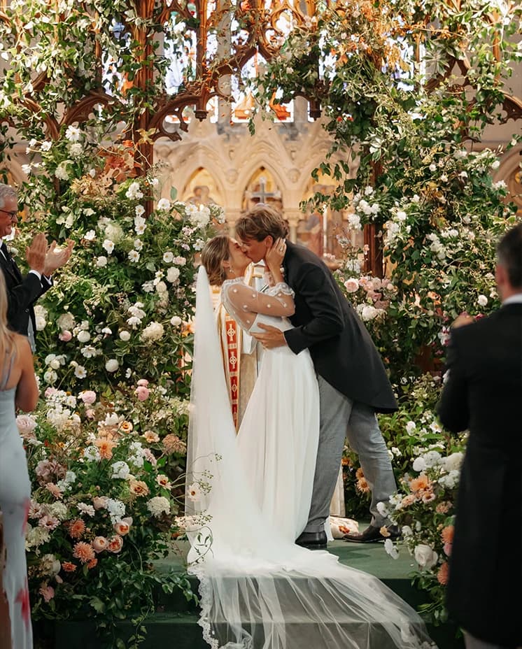 Bride and groom sharing their first kiss beneath a breathtaking floral ceremony installation inside a historic church.