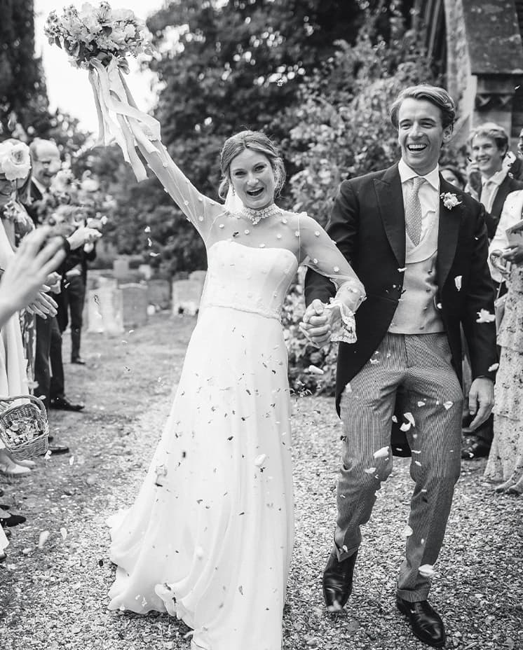 Newlyweds walking through a confetti shower after their church wedding, with the bride raising her bouquet in celebration.