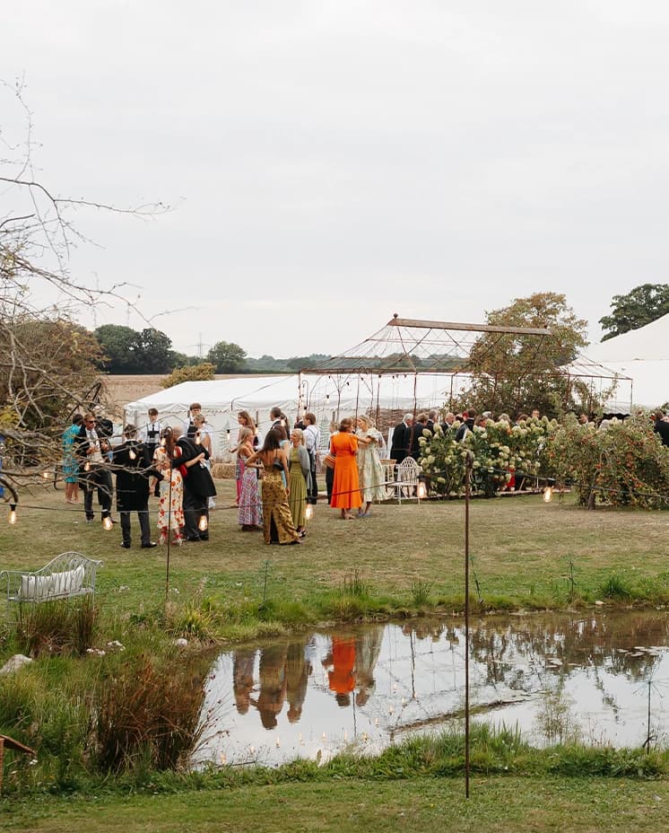 Wedding guests mingling beside a luxury marquee and garden lounge area overlooking a pond during a countryside wedding reception.