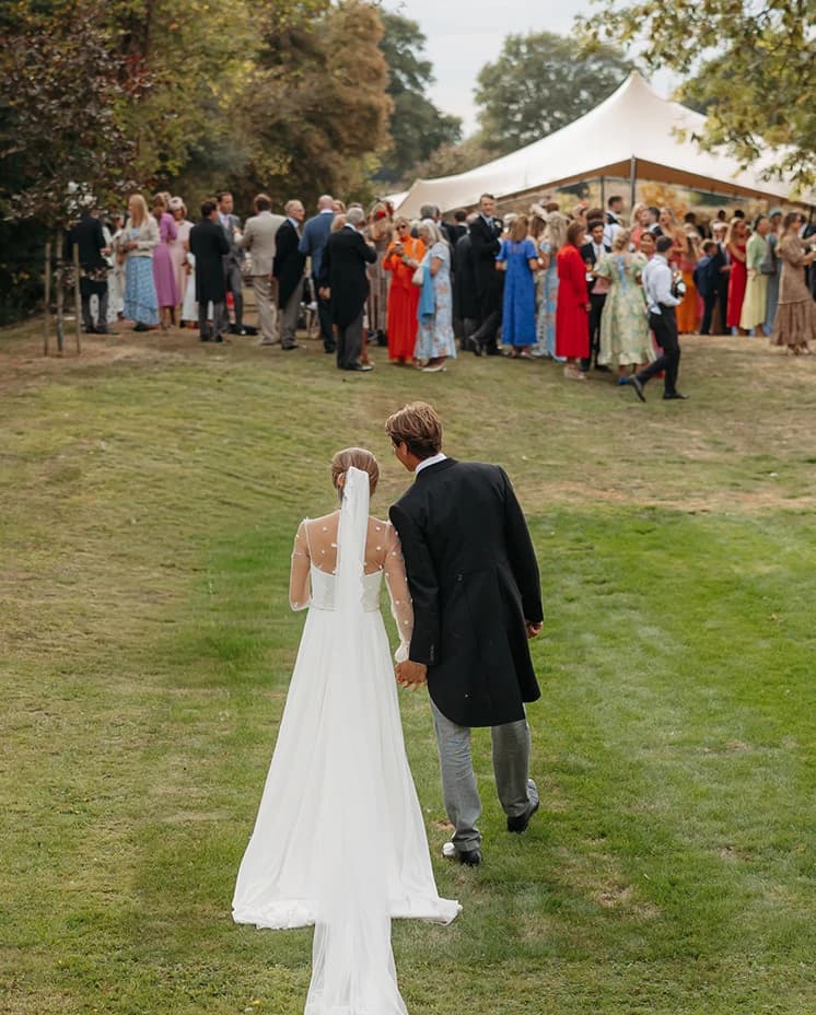Bride and groom walking hand in hand across the lawn towards their guests and marquee reception at a countryside wedding.