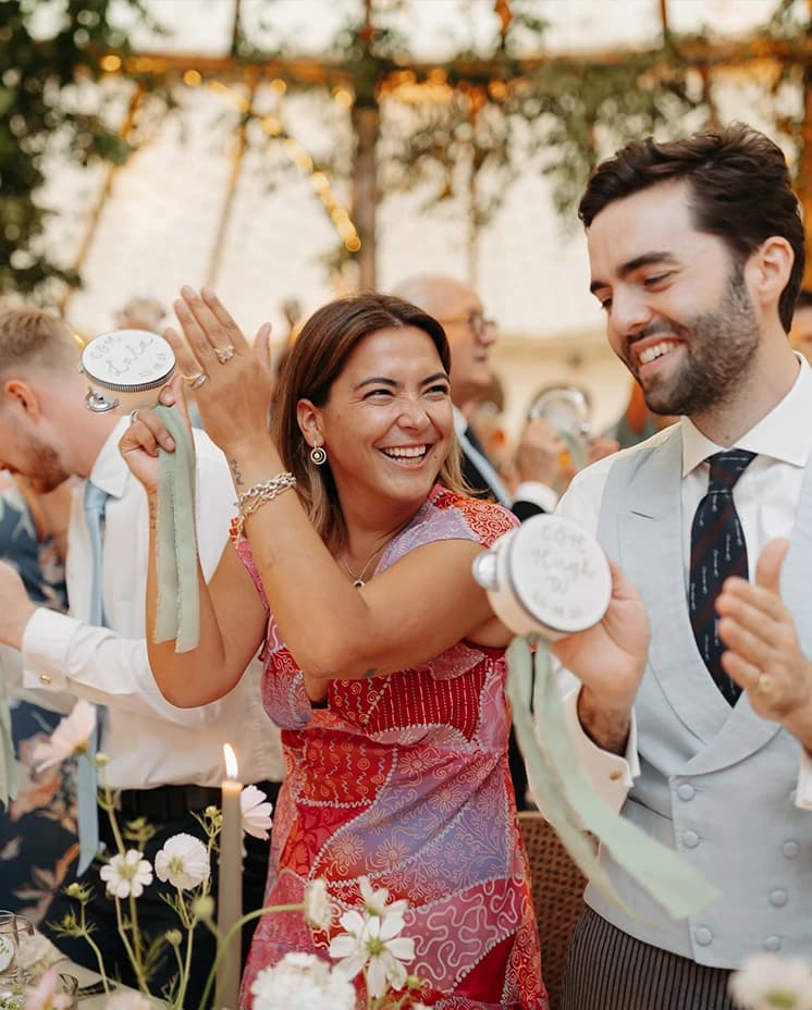Wedding guests playing personalised tambourines and celebrating at a marquee reception beneath cascading greenery and floral décor.