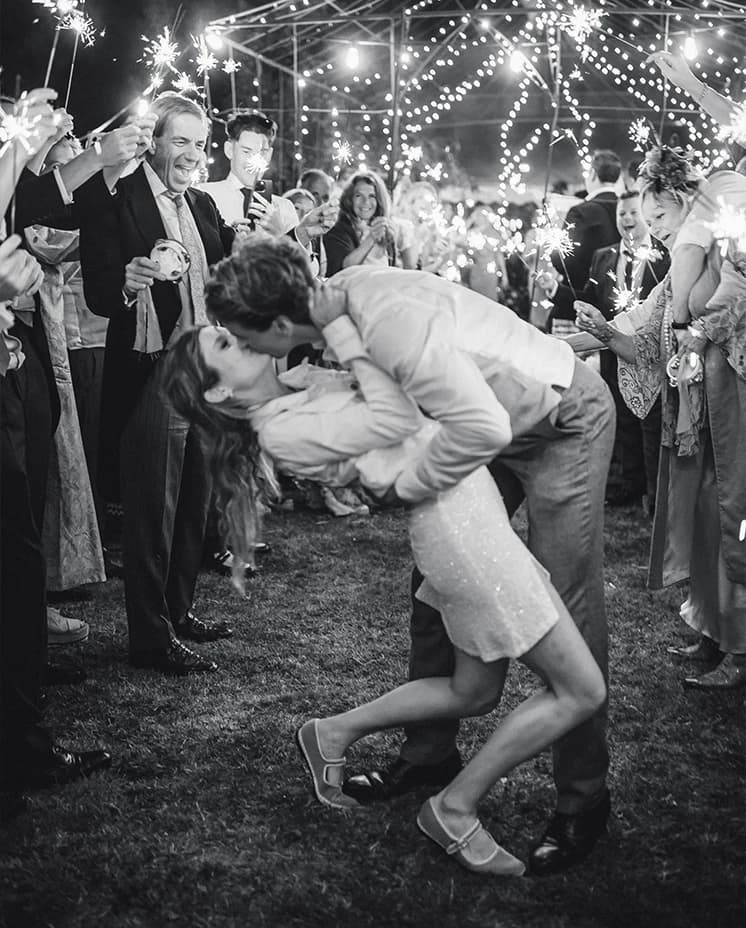 Bride and groom sharing a romantic kiss beneath sparklers and twinkling lights, surrounded by guests during an evening countryside wedding celebration
