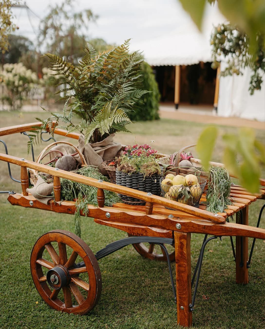 Rustic wooden cart decorated with seasonal produce, herbs and greenery outside a marquee wedding in the countryside.