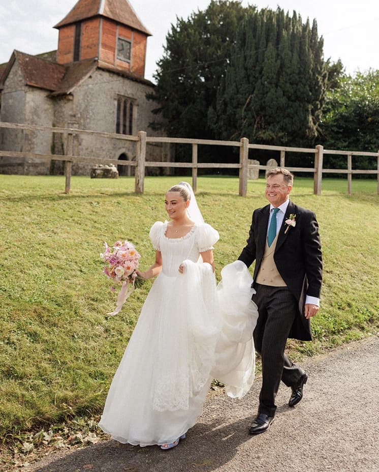 Bride walking with her father outside a historic English countryside church before the wedding ceremony, wearing a white puff-sleeve wedding dress, veil and carrying a pastel bridal bouquet. 