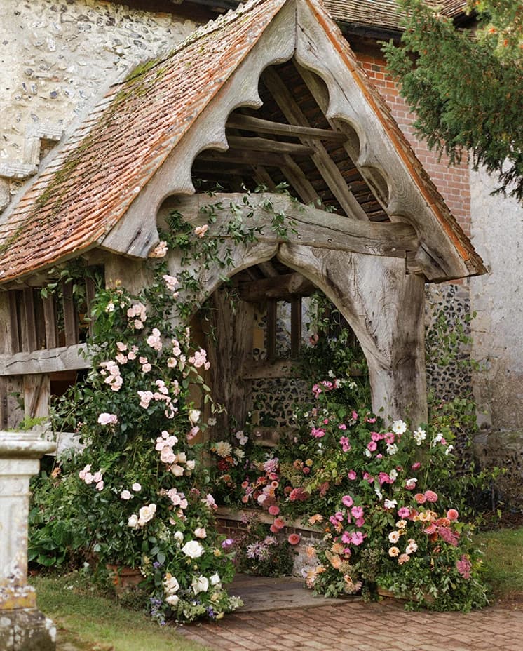 Historic English church lychgate adorned with abundant pink and white floral arrangements, creating a romantic entrance for a countryside wedding ceremony.
