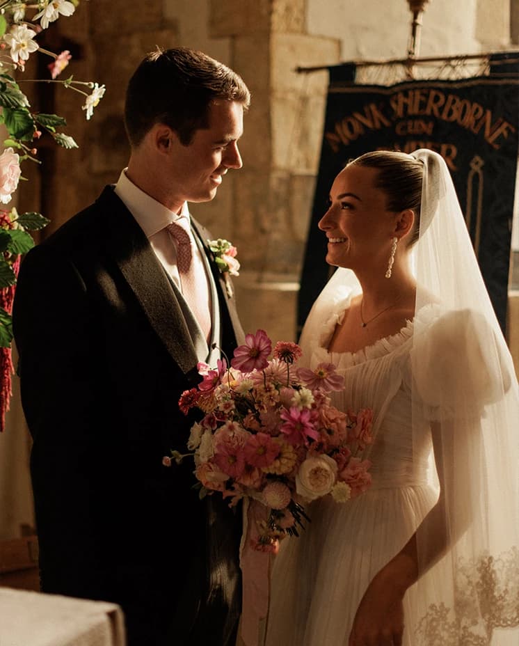 Bride and groom sharing a smile inside a historic English church, with the bride holding a pink and blush wedding bouquet during their romantic wedding ceremony.