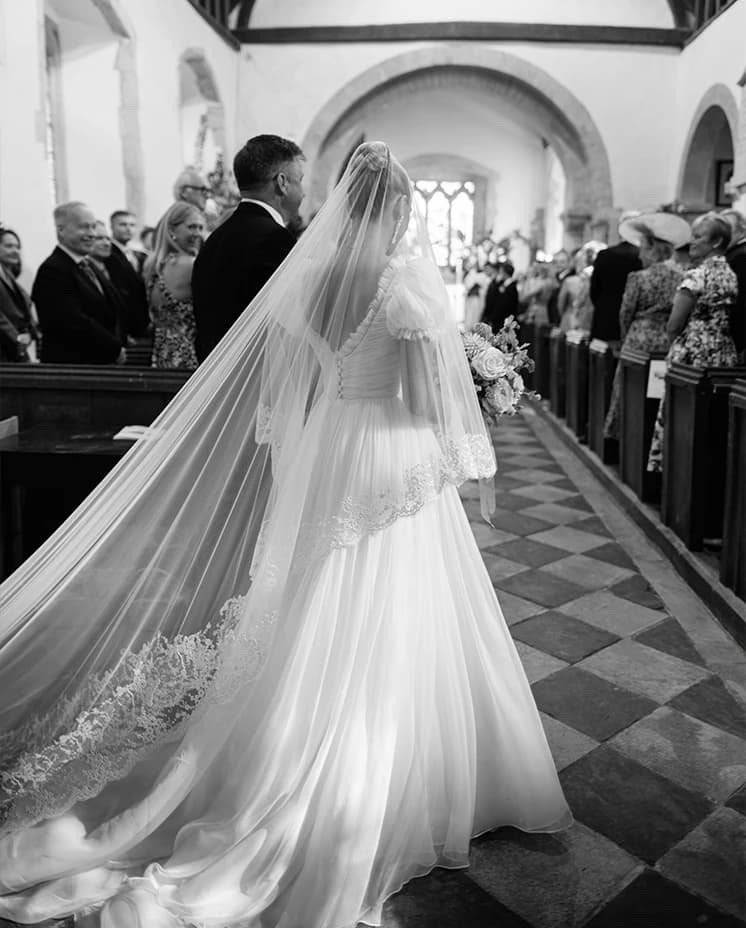 Bride walking down the aisle in a historic English church, wearing a flowing lace-edged veil and elegant wedding dress during a romantic church wedding ceremony.