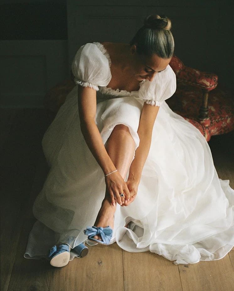 Bride fastening her blue  Loeffler Randall wedding shoes while getting ready, wearing a romantic puff-sleeve wedding dress before her English countryside wedding ceremony.