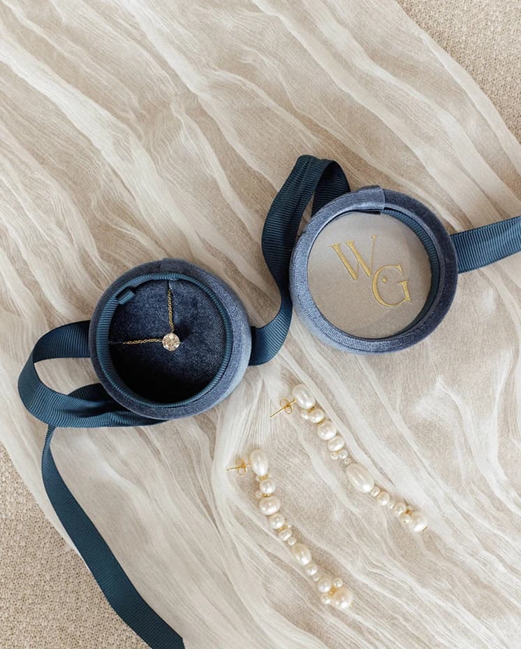 Flat lay of bridal accessories featuring pearl drop earrings, a diamond necklace and velvet ring boxes on a soft wedding veil, styled for an elegant wedding morning.