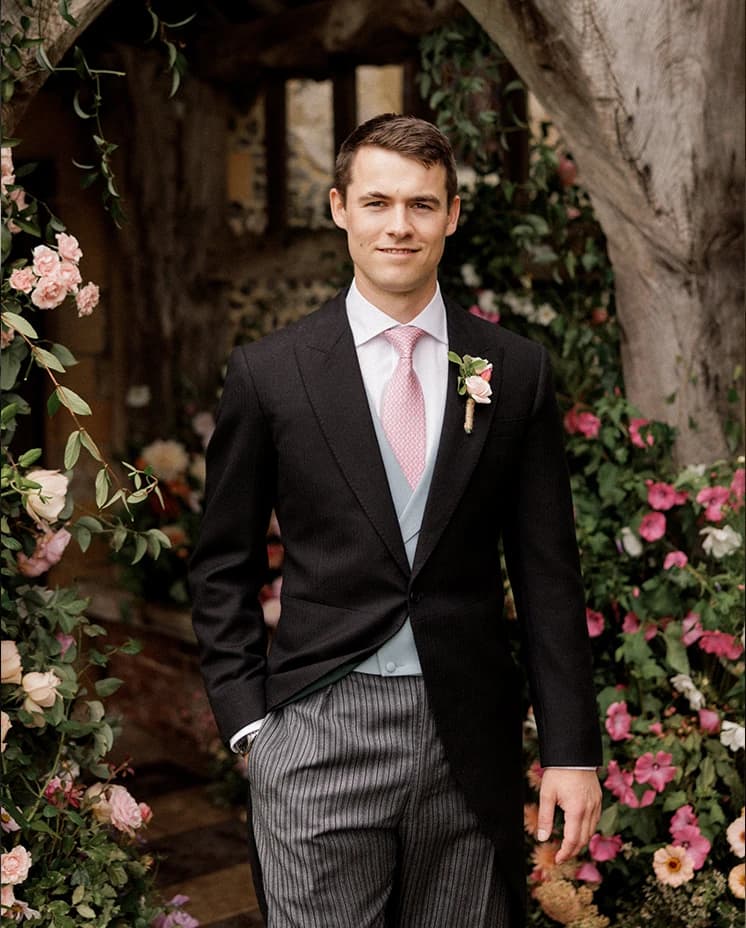 Groom in a Buckleigh of London classic morning suit with a pink tie and boutonnière, standing beneath a flower-filled church lychgate before an English countryside wedding ceremony.
