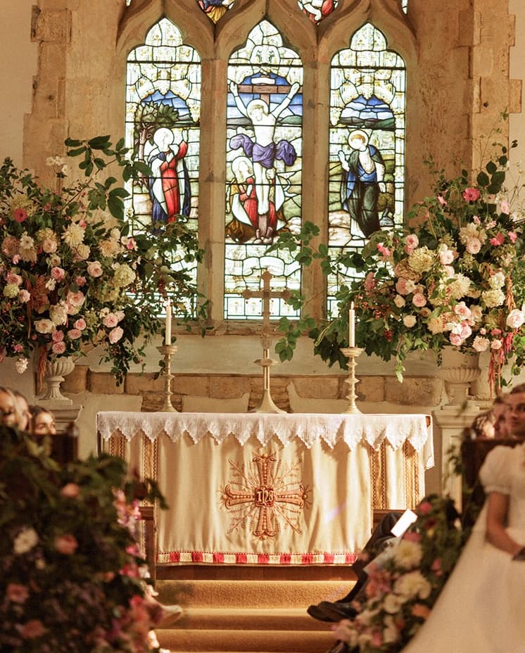 Historic English church altar adorned with abundant pink and white floral arrangements beneath stained glass windows, beautifully styled for a romantic countryside wedding ceremony. 