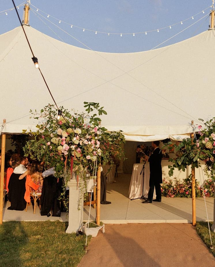 Sailcloth wedding marquee adorned with abundant floral arrangements and festoon lighting, creating a romantic English countryside wedding reception at sunset. 