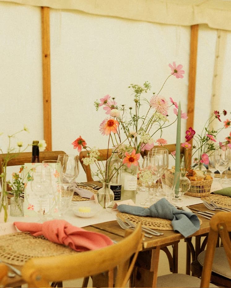 Elegant wedding reception table styled with colourful seasonal flowers, pastel candles and rustic place settings inside a sailcloth marquee at an English countryside wedding. 