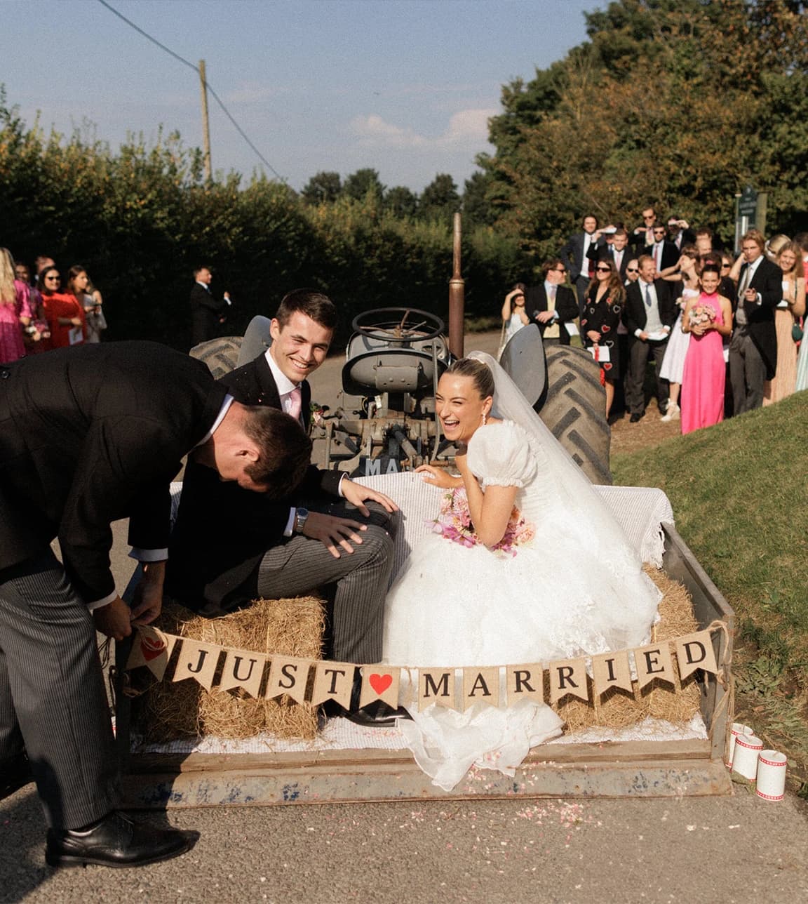 Bride and groom celebrating in a vintage tractor trailer with a "Just Married" sign, laughing with wedding guests after their English countryside wedding ceremony.
