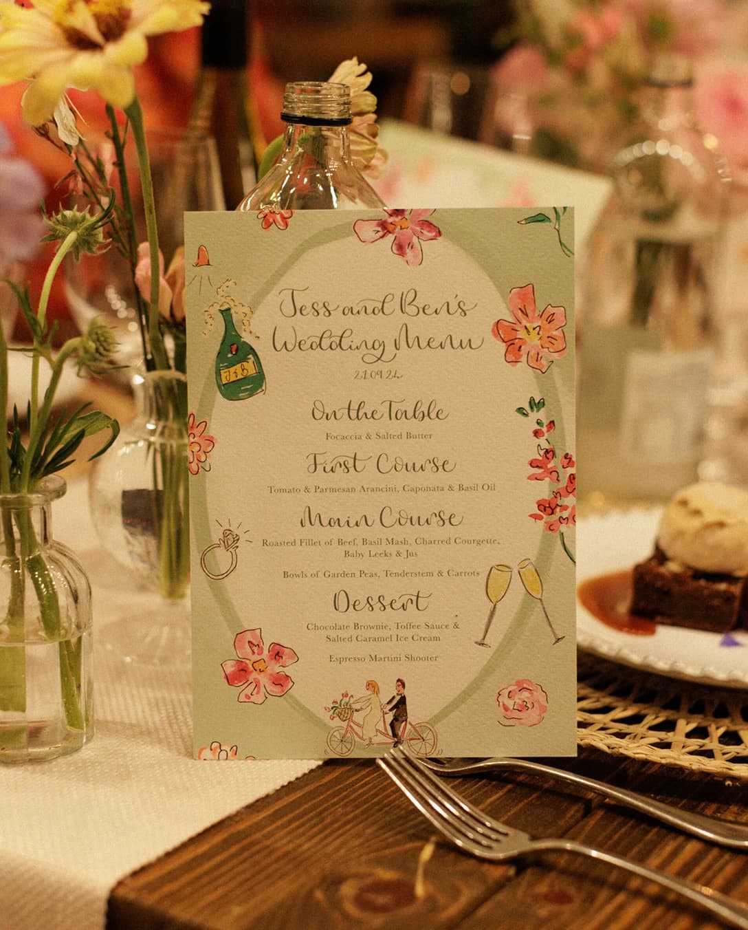 Illustrated wedding menu on a beautifully styled reception table with seasonal flowers and elegant place settings inside a sailcloth marquee at an English countryside wedding.