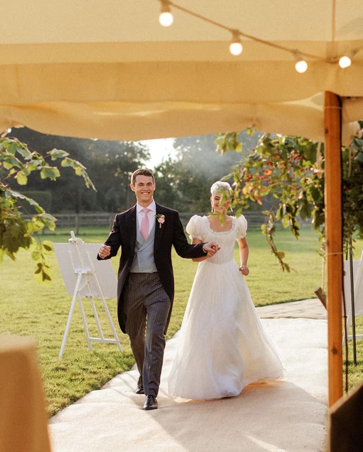 Bride and groom making their grand entrance into a sailcloth marquee, celebrating their English countryside wedding reception beneath festoon lights.