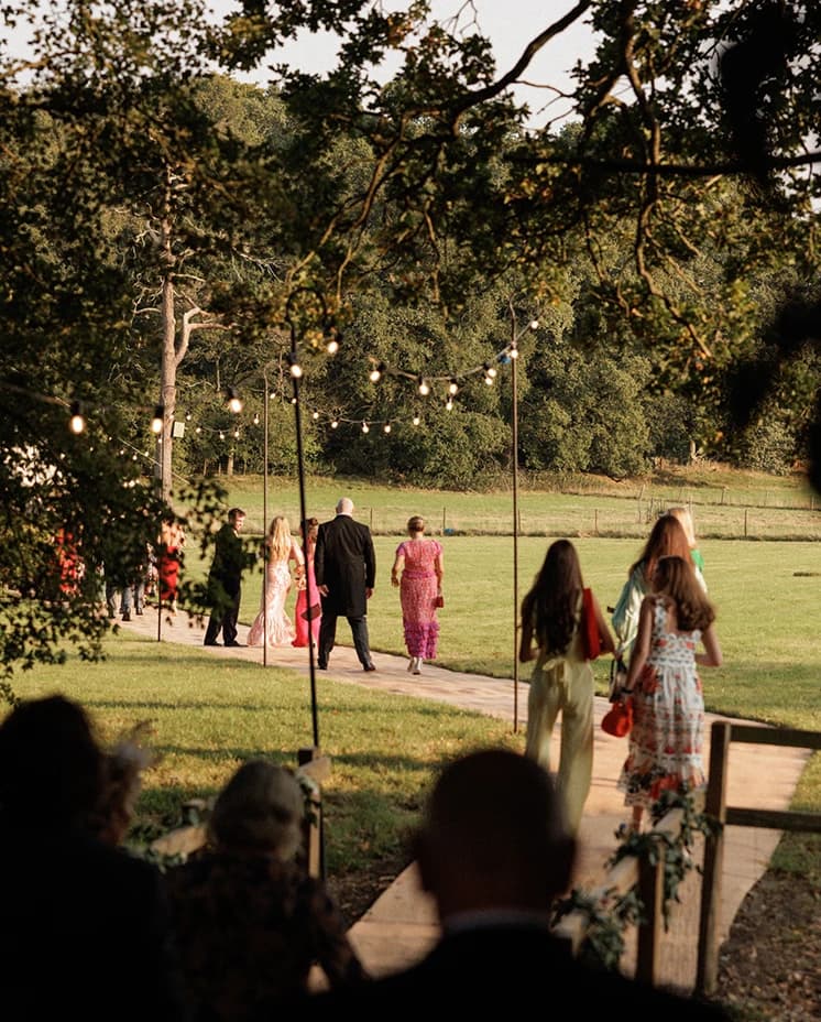 Wedding guests strolling beneath festoon lights towards a sailcloth marquee, capturing the relaxed atmosphere of an English countryside wedding reception.